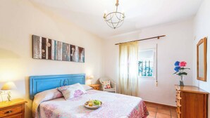 A bedroom with a blue headboard, a bedside table with a lamp, a dresser, and a window with curtains. - Villa Carmela Nerja (Nerja)