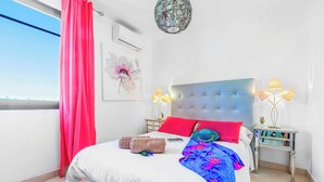 Quarto com cama, almofadas, mesa de cabeceira, abajur e janela com cortinas.
