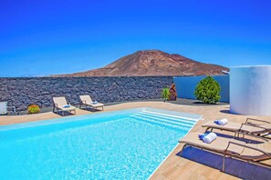 A swimming pool with lounge chairs, a stone wall, and a mountain in the background. - Villa Esmeralda (Yaiza)