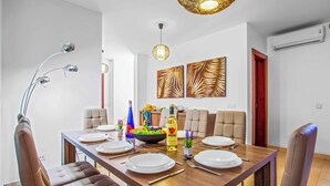 Uma sala de jantar com uma mesa de madeira posta para seis pessoas, rodeada por cadeiras de couro. Há duas luminárias fixadas na parede e um lustre pendente.
