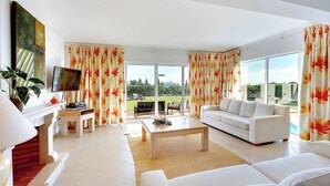 A modern living room with a sofa, a coffee table, and a TV. There is a large window with curtains and a view of greenery outside.