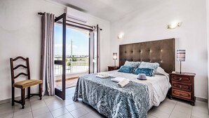 A bedroom with a large bed, a wooden nightstand, a chair, and a view through a glass door. - Villa Canto (Albufeira)