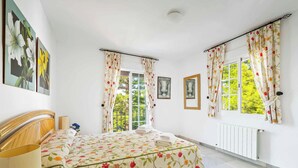 A bedroom with a bed, two windows with curtains, a radiator, and artwork on the walls. - Villa Clarence (Nerja)