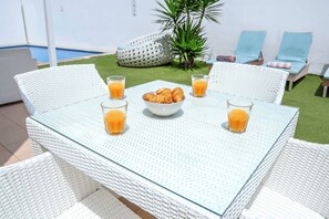 Una mesa con un bol de croissants y vasos de jugo de naranja, rodeada de sillas de mimbre blanco.
