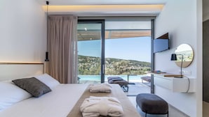A modern hotel room with a large bed, a wooden bedside table, a bench, and a view of a mountainous landscape through a sliding glass door. - Villa Beyond the Horizon (Lefkada)