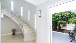Eine Treppe mit Glasgeländer führt zu einer Zwischenebene, einer Tür zu einem Außenbereich mit Baum und Bank sowie einer Wandleuchte.