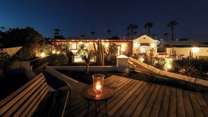A wooden deck with a glass table holding a candle, surrounded by plants and a pool. - Villa Dos Piedras (Tías)