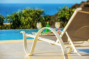 A lounge chair by a pool with a table set for two, glasses, and a bottle.