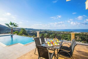A terrace with a pool, dining table, chairs, and a scenic view of the sea and hills.