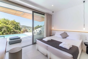 A modern hotel room with a large bed, a bench, and a view of a pool and greenery. - Villa Beyond the Dream (Lefkada)