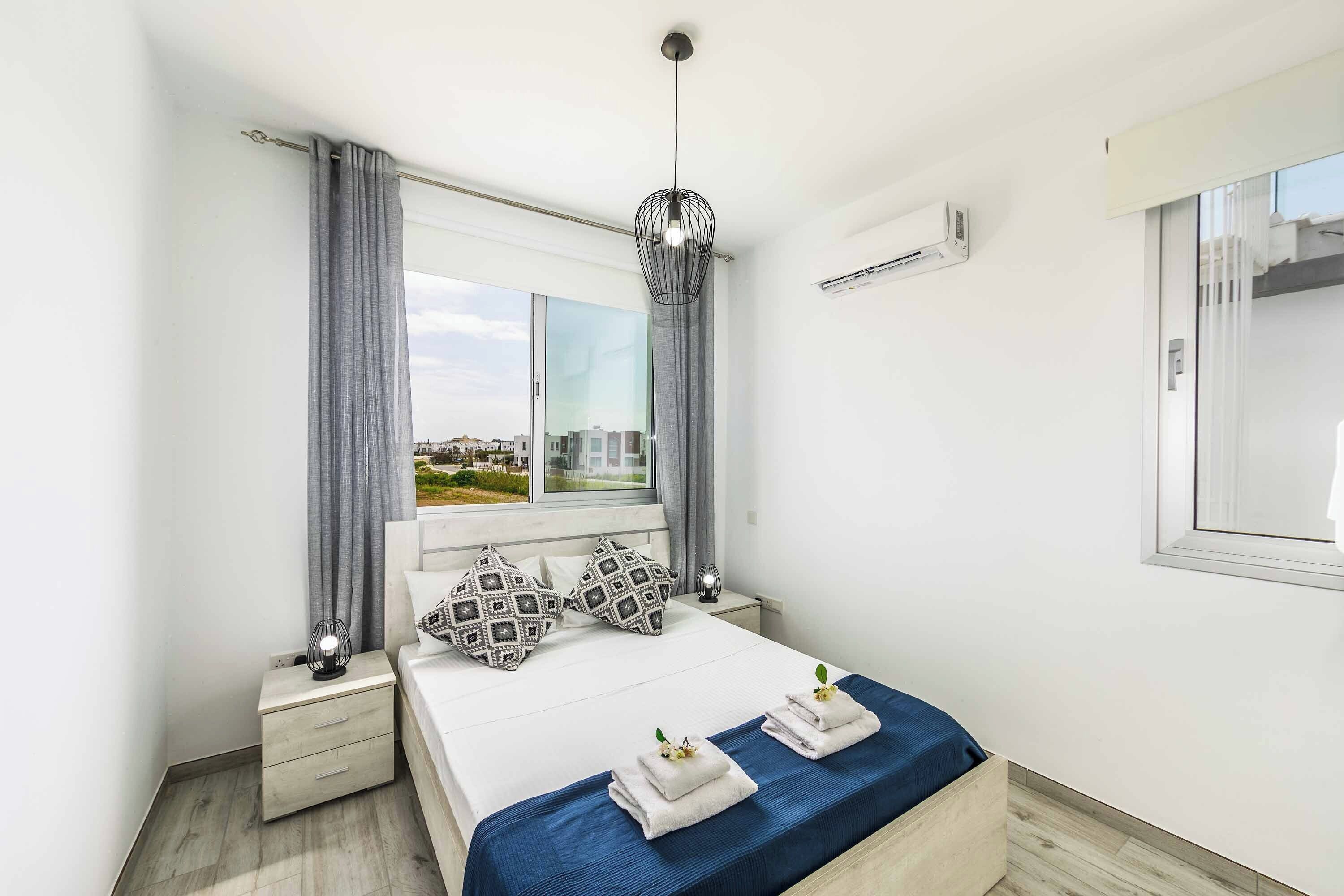 A modern bedroom with a large bed, grey curtains, a hanging light fixture, and a view of buildings outside.