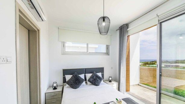 A modern bedroom with a large bed, a bedside table, a lamp, and a view of the outdoors. - Villa Alasia (Paralimni)