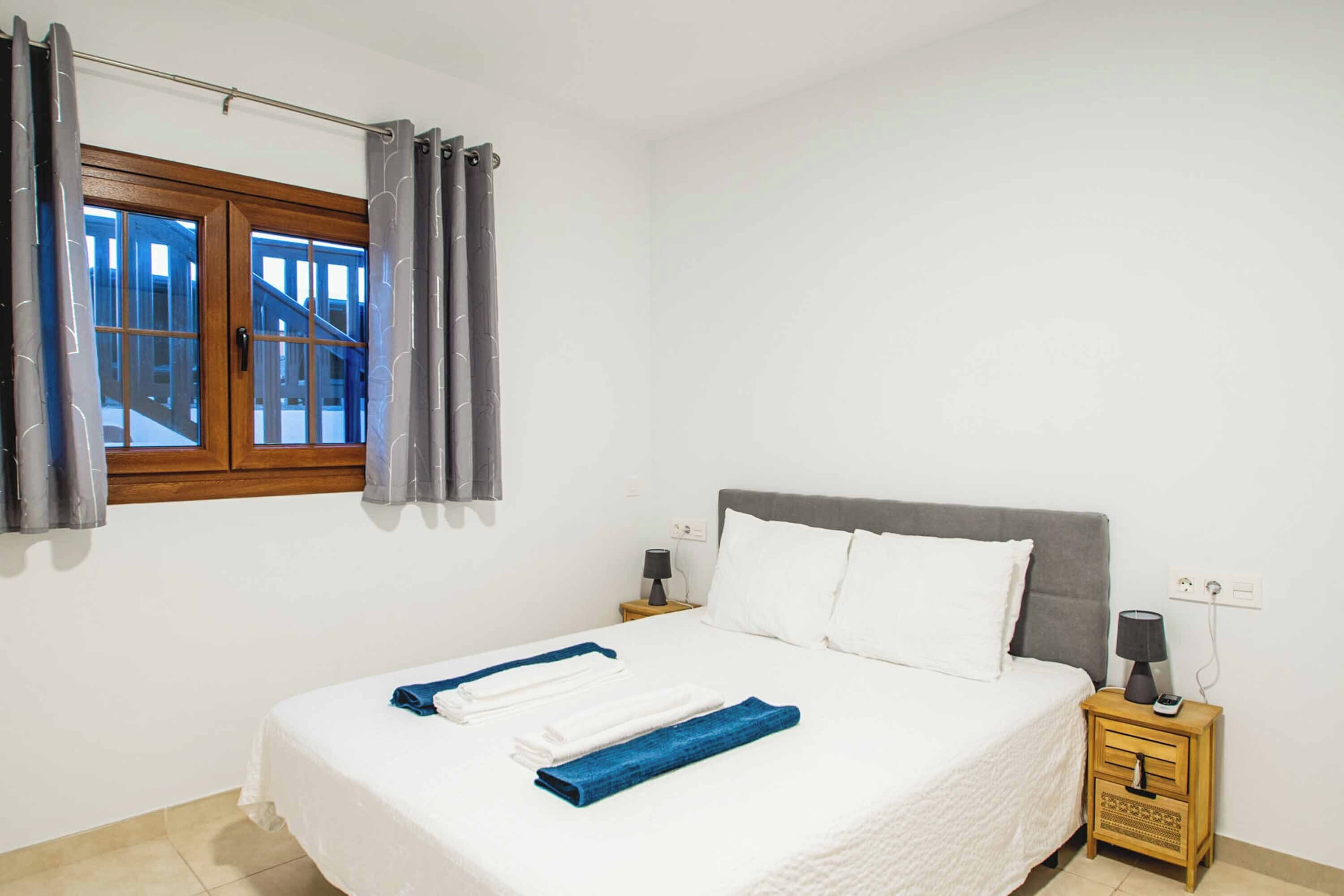 Ein Schlafzimmer mit einem Bett, weißer Bettwäsche, einem Nachttisch mit Lampe und einem Fenster mit Vorhängen.