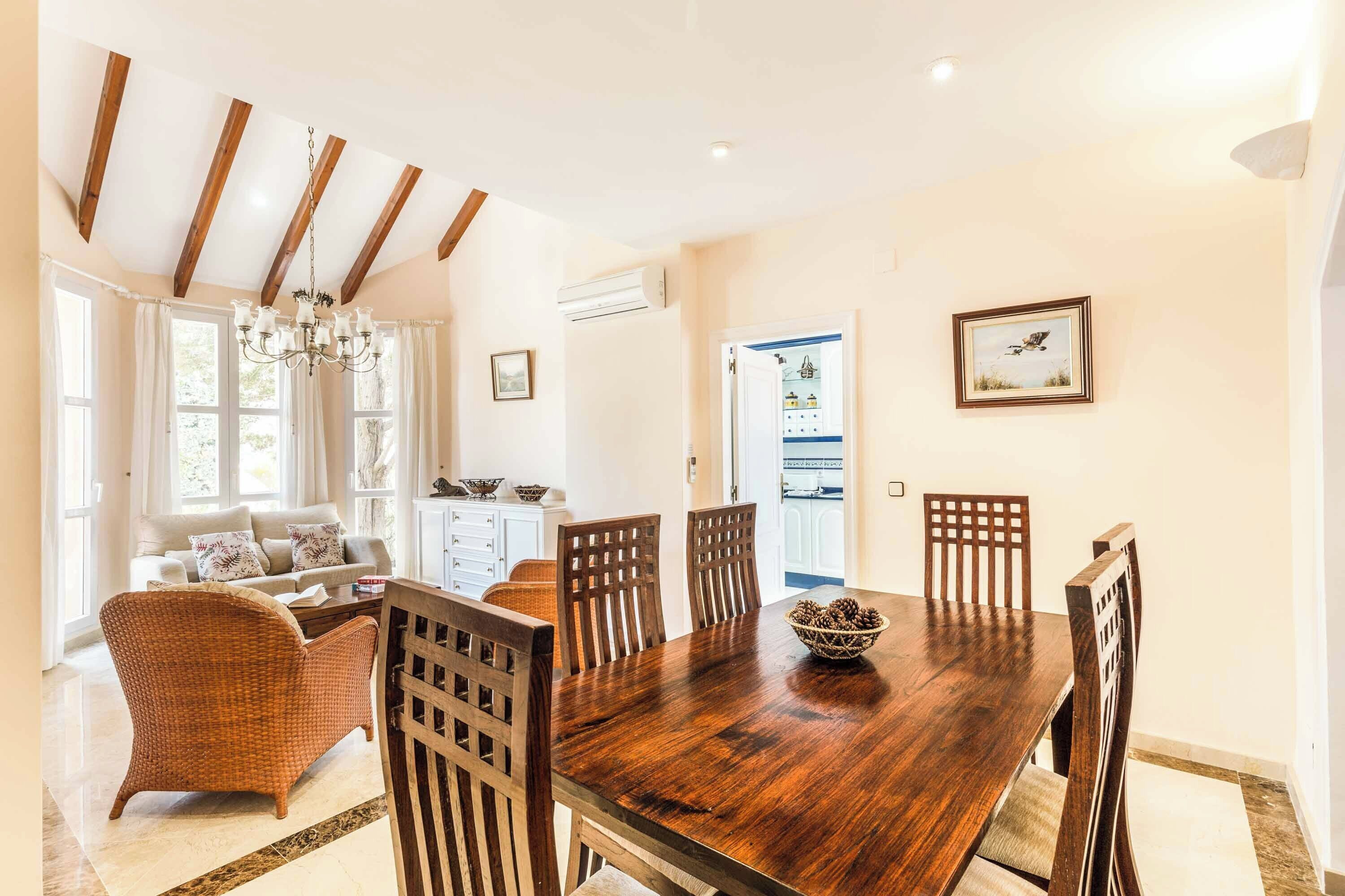 A dining area with a wooden table and chairs, a chandelier, and a view of the kitchen through an open door.