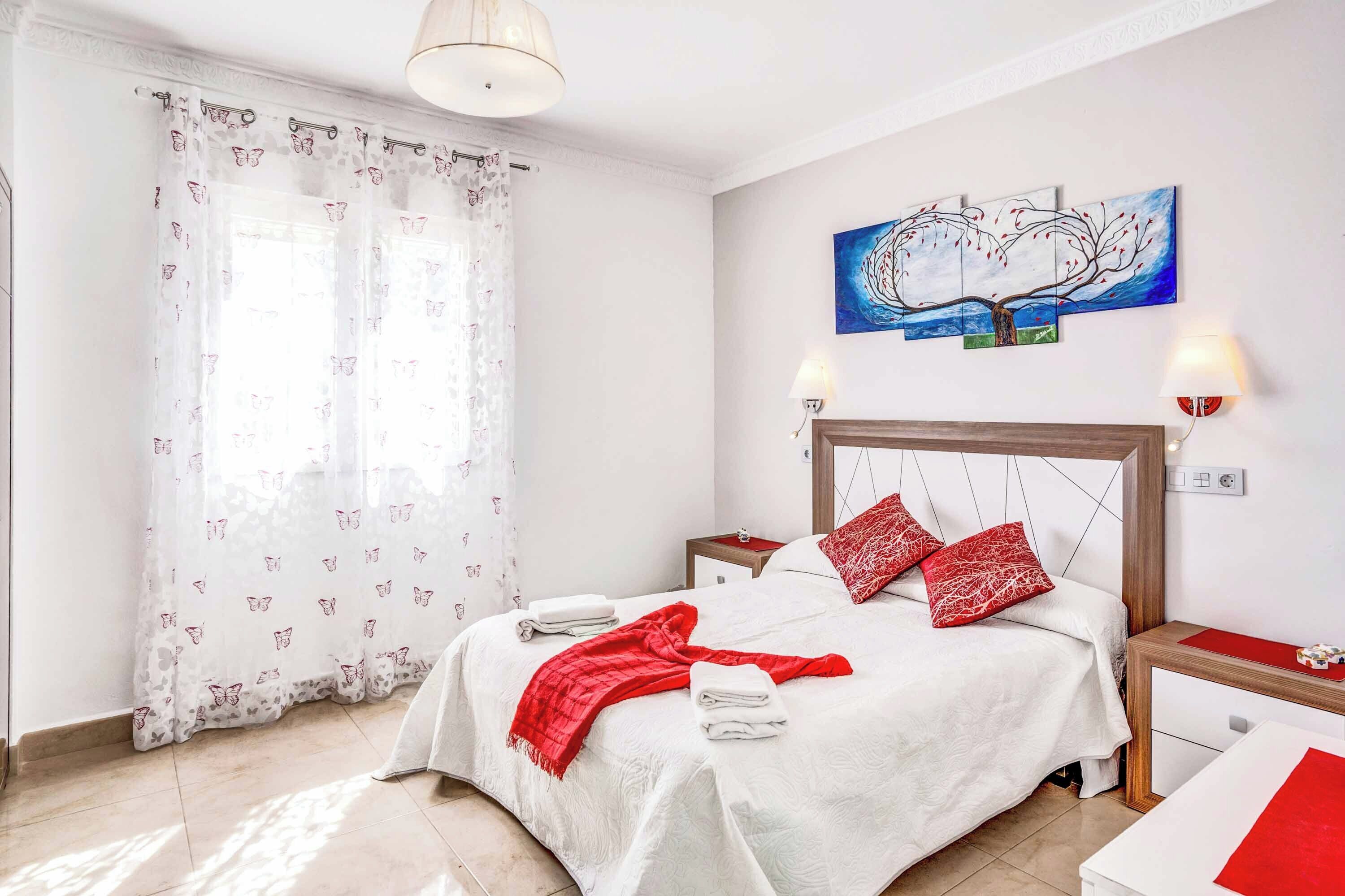 Une chambre à coucher comprenant un lit, une table de chevet, une commode, un tableau au mur et une fenêtre avec des rideaux.