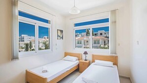 Ein helles, modernes Schlafzimmer mit zwei Einzelbetten, einem kleinen Nachttisch mit Lampe und großen Fenstern mit Blick auf die Stadt.