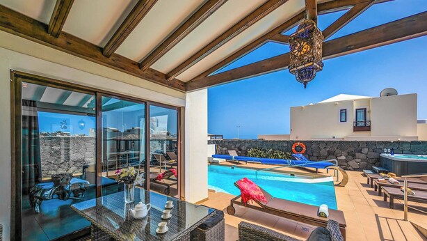 A sunlit outdoor area with a pool, lounge chairs, and a glass-enclosed dining area. - Villa Xian (Yaiza)