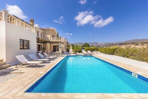 A swimming pool with lounge chairs, a stone building with a balcony, and a view of the hills. - Villa Cleco (Pegeia)