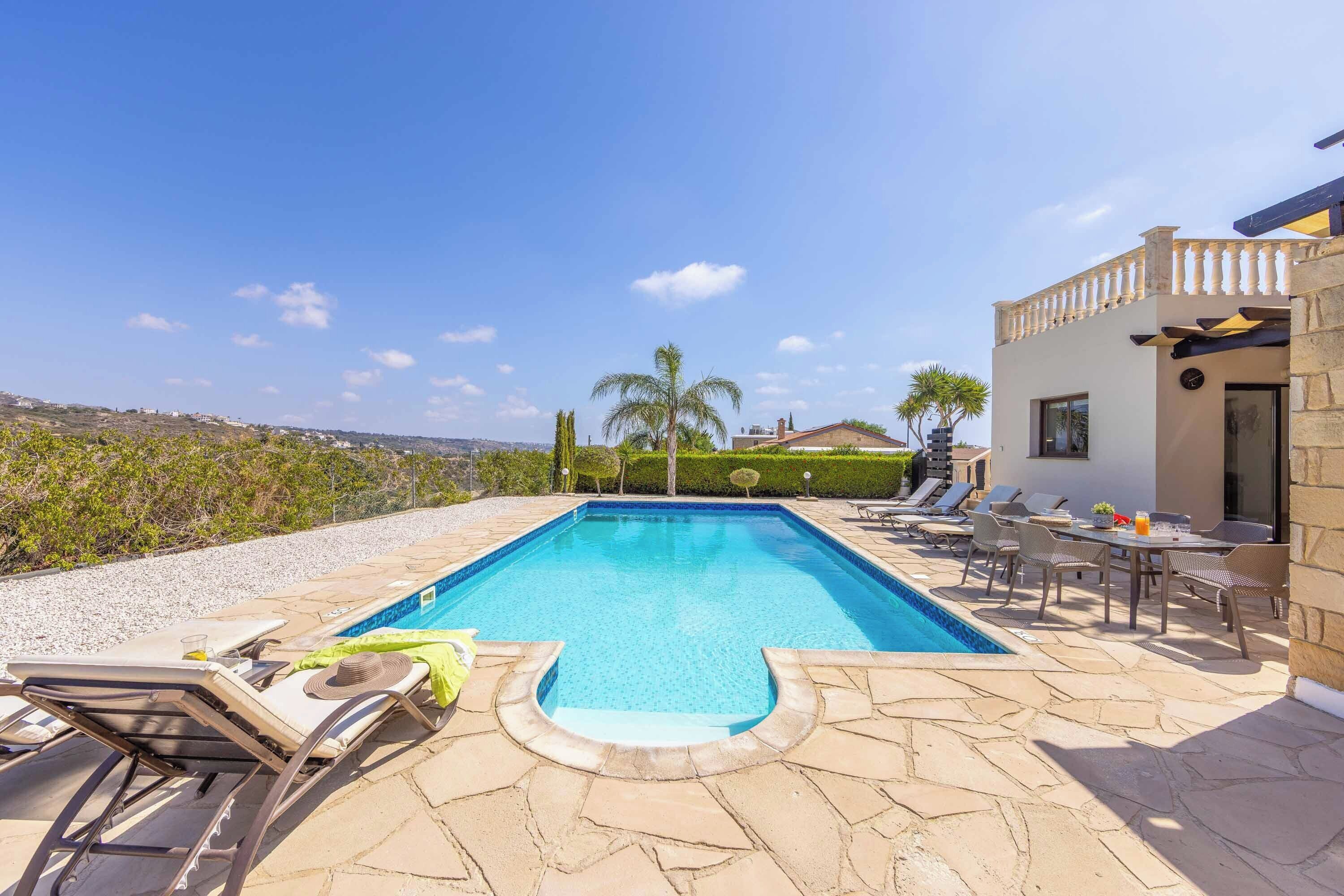 Une piscine rectangulaire bordée d’une terrasse en pierre, entourée de chaises longues et d’une table avec des chaises.