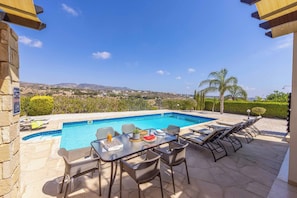 A poolside area with a table, chairs, and lounge chairs. - Villa Cleco (Pegeia)