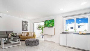 A modern living room with a grey sofa, a glass coffee table, and a grey ottoman. There is a kitchen area with white cabinets and a window showing a clear sky. - Villa Mi Deseo (Tías)