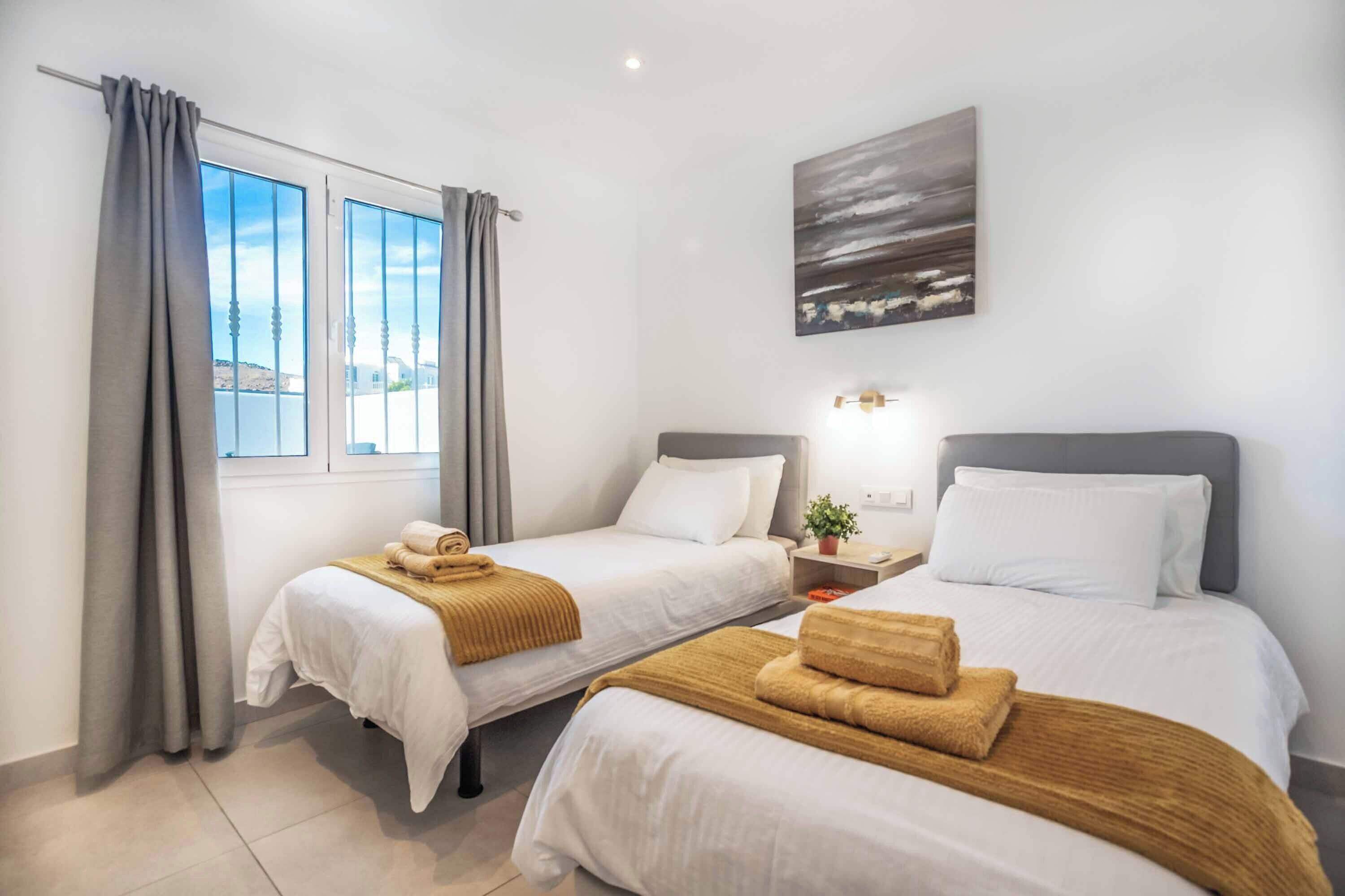 A hotel room with two beds, a window with curtains, a painting on the wall, and a bedside table with a lamp.