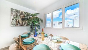 A dining area with a wooden table set for six, surrounded by chairs, with a Buddha painting on the wall and a large window offering a view of buildings. - Villa Mi Deseo (Tías)