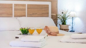 Una cama bien hecha con sábanas blancas, una bandeja con dos vasos de líquido amarillo, una planta pequeña en maceta y una toalla doblada.