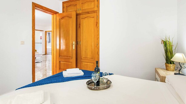 A hotel room with a bed, a bedside table with a lamp, a tray with a bottle and glasses, a potted plant, and a wooden wardrobe. - Villa Enzo Moraira (Teulada)