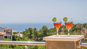 Deux verres d’une boisson rouge, décorés d’une tranche de citron vert, sur un balcon offrant une vue sur la mer et la verdure.