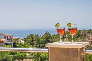 Two glasses of red beverage with lime garnish on a balcony with a view of the sea and greenery.