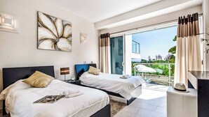 A modern hotel room with two beds, a large window with curtains, a balcony with outdoor furniture, and a view of greenery.