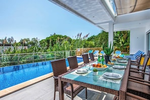 A dining area with a glass table and wicker chairs, overlooking a swimming pool and lush greenery.