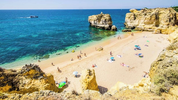 A beach with clear turquoise water, rocky formations, and people sunbathing.