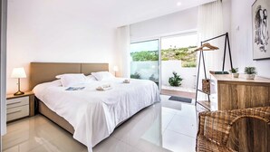A modern bedroom with a large bed, a nightstand, a lamp, a wicker chair, a wooden dresser, and a view of greenery outside. - Villa Aventura (Albufeira)