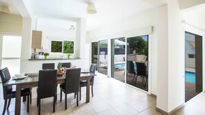 Un comedor moderno con una mesa dispuesta para cuatro personas, una cocina al fondo y una piscina visible a través de puertas de vidrio.