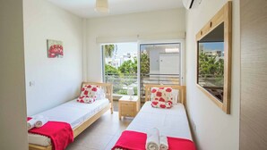 Habitación de hotel con dos camas individuales, una mesita, una silla, un ventanal con vistas a edificios y vegetación, y un balcón.
