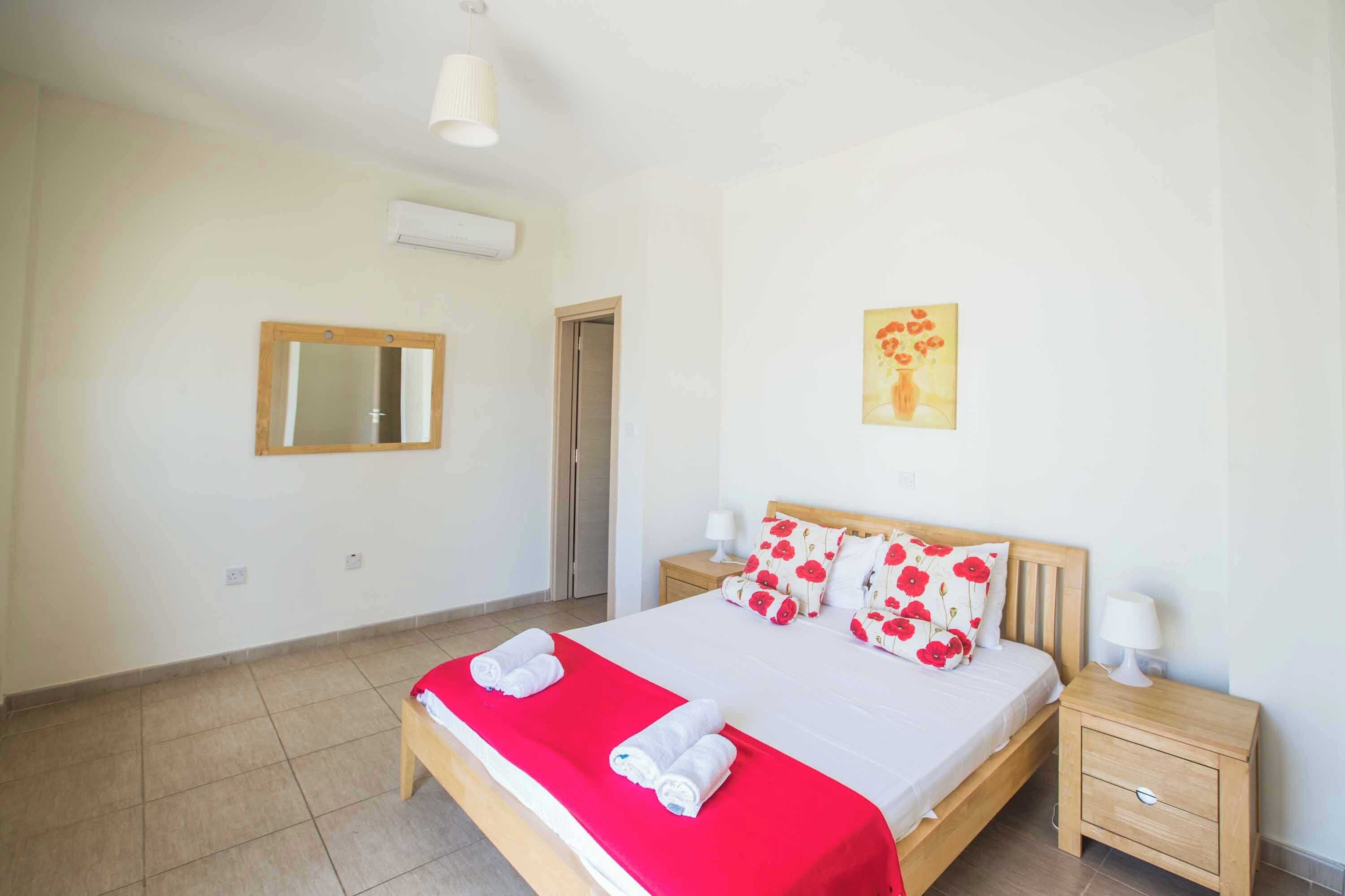 A bedroom with a bed, two bedside tables, a nightstand, a mirror, and a wall-mounted air conditioner.