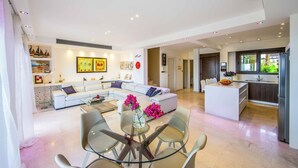 A modern living room with a glass dining table, white sofas, and a kitchen area with a central island. - Villa El Greco (Paralimni)