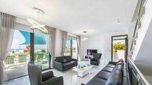 A modern living room with a sofa set, a coffee table, and a TV. Large windows offer a view of the outdoors. - Villa Alderbaran (Pegeia)