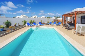 A swimming pool with blue water, surrounded by blue lounge chairs and a wooden pergola. - Villa Luar (Yaiza)