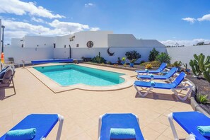A pool area with blue lounge chairs, a pool, and a white building with a crescent moon design. - Villa Luar (Yaiza)