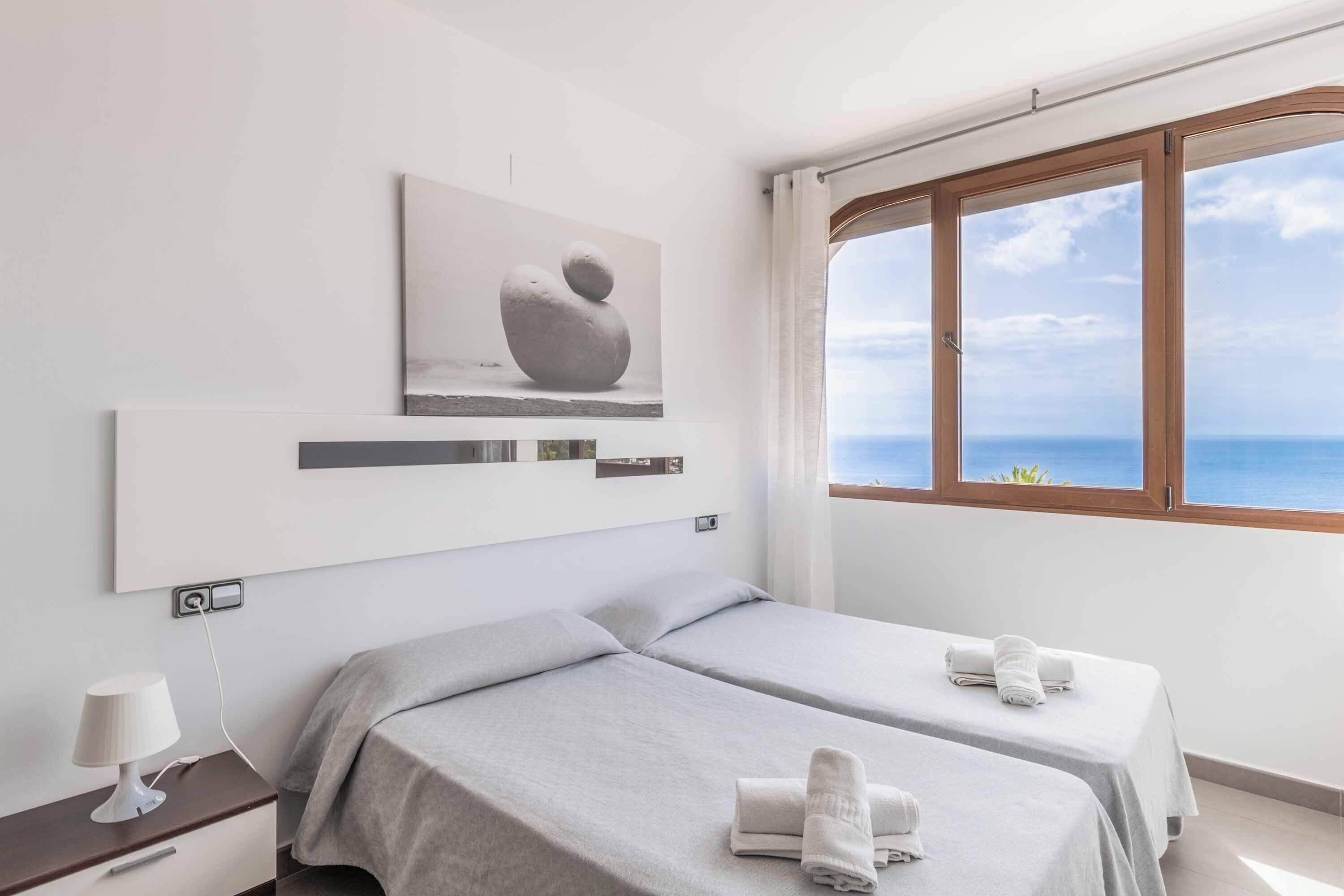 Un hotel moderno con dos camas, una mesita de noche con lámpara, un ventanal con vistas al mar y una obra de arte minimalista en la pared.