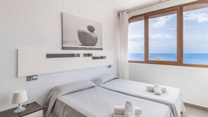 Un hotel moderno con dos camas, una mesita de noche con lámpara, un ventanal con vistas al mar y una obra de arte minimalista en la pared.