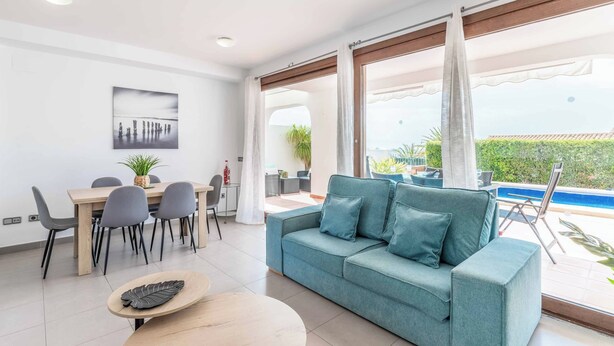 Una sala de estar moderna con sofá, mesa de comedor y sillas. Hay un ventanal con cortinas, una piscina visible al exterior y una planta en la mesa.
