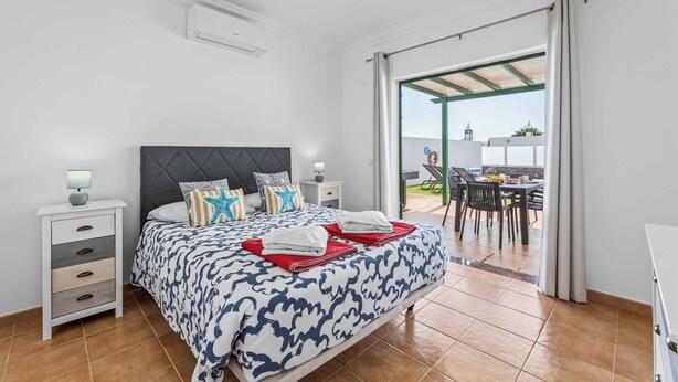 A bedroom with a bed, bedside tables, and a view of an outdoor dining area. - Villa Bonaci (Yaiza)