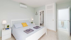 Un dormitorio con una cama, mesitas de noche, una lámpara de noche, aire acondicionado montado en la pared y una puerta que da a un baño.