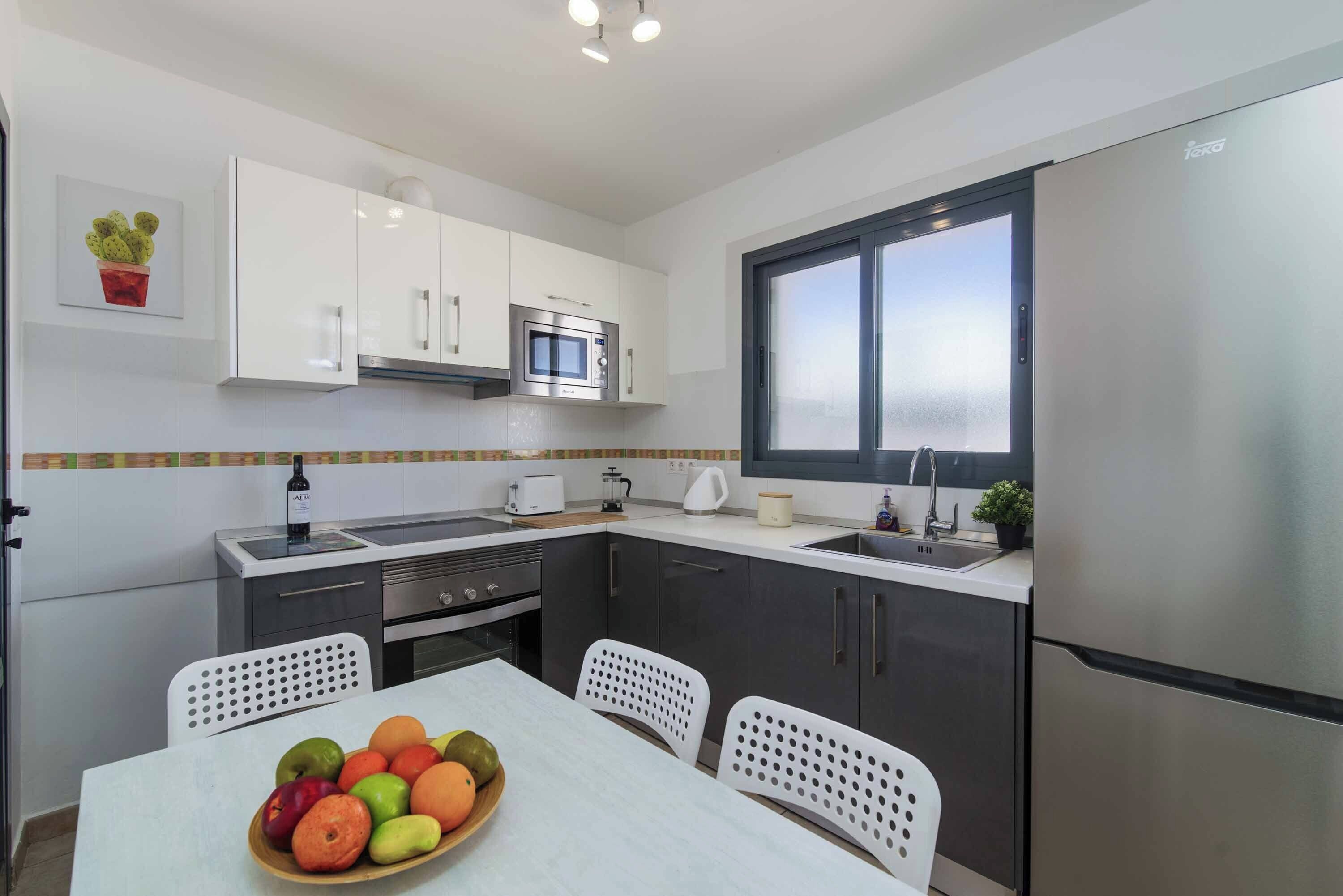 Una cocina moderna con armarios blancos, un refrigerador de acero inoxidable, un microondas y un lavabo. Hay una mesa de comedor con sillas y un tazón de frutas.