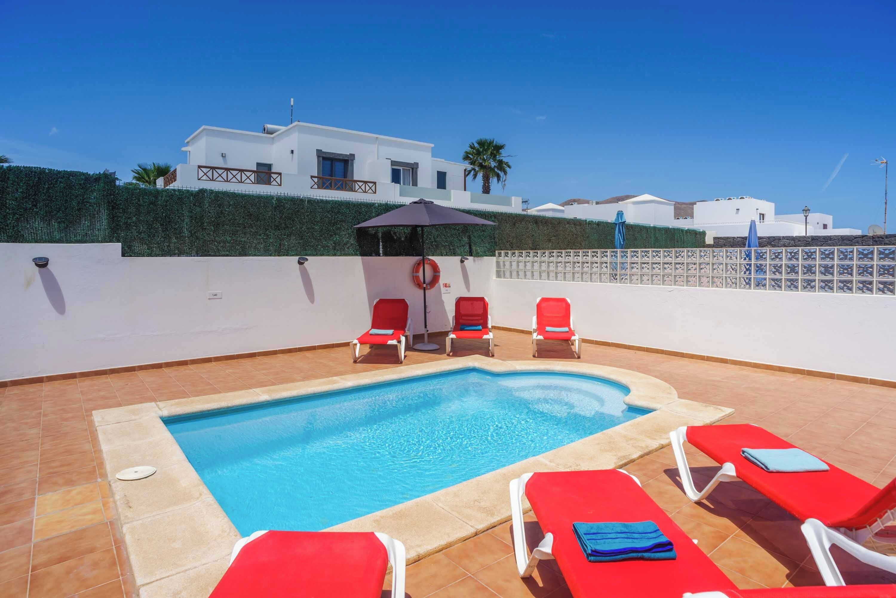 A swimming pool with red lounge chairs and a covered seating area.