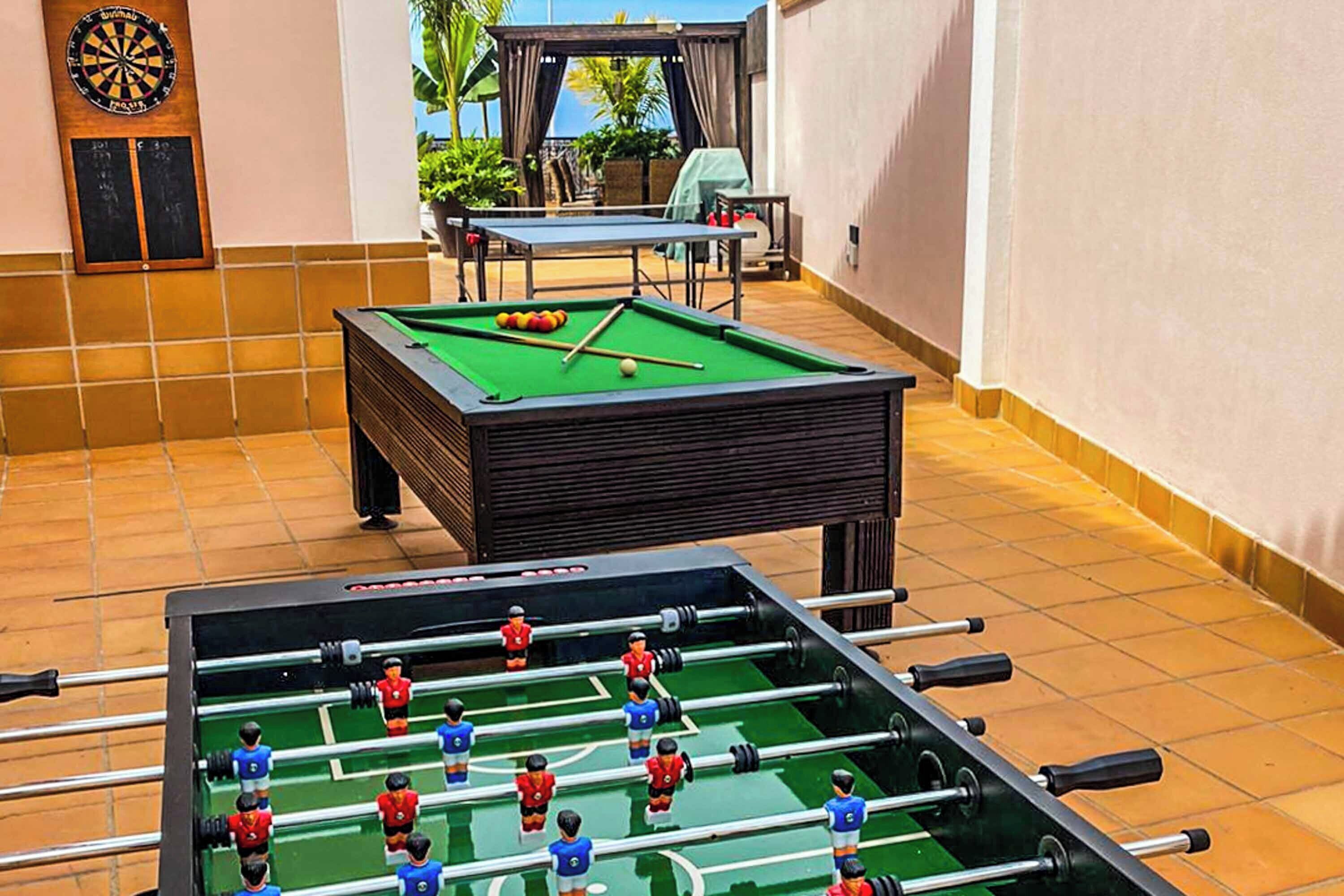 Ein Billardtisch mit einem Kicker davor, eine Dartscheibe und eine Terrassenfläche mit Tisch und Stühlen.